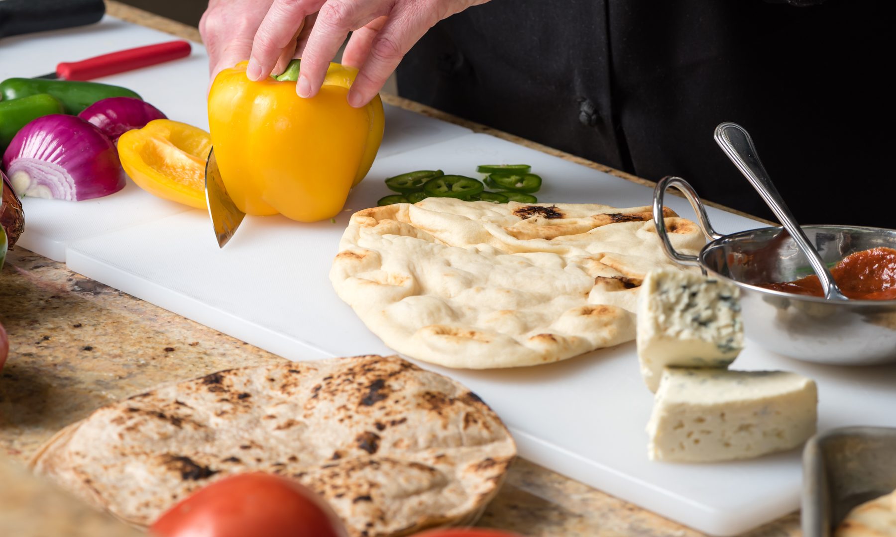 Chef preparing a meal by slicing a pepper. Stonefire Original Naan is on the cutting board with vegetables, cheese and sauces nearby in restaurant preparation style.